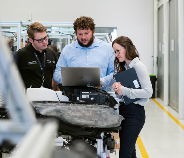 Three people working in a machine shop standing on a laptop Three people working in a machine shop standing on a laptop