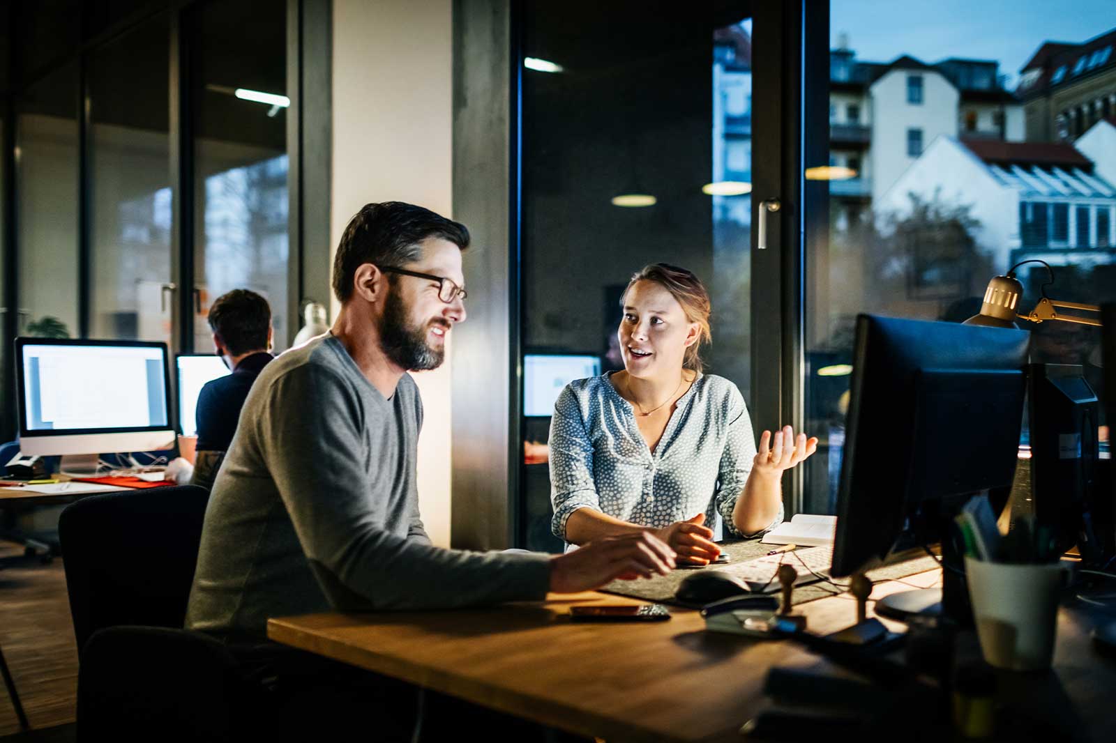 Two people talking in front of a PC with evening atmosphere Two people talking in front of a PC with evening atmosphere