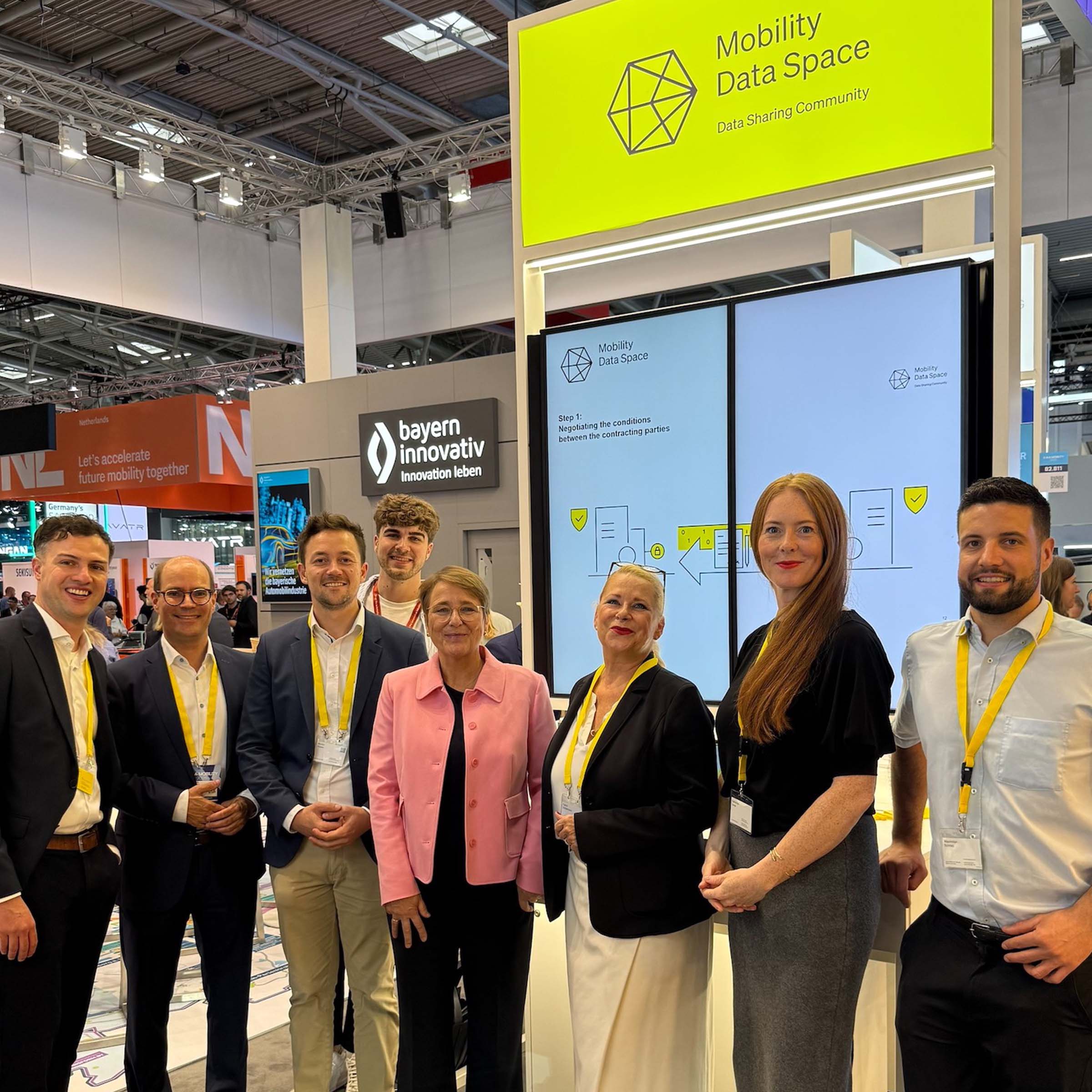 Eight people are standing in front of a screen in a trade fair hall, smiling at the camera. In the background, other visitors and exhibition stands can be seen.