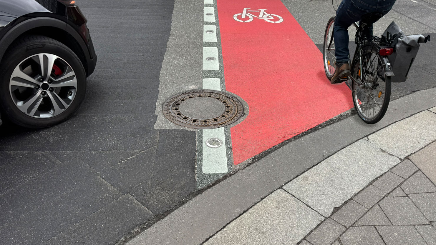 A cyclist is riding on a cycle path marked in red with a dashed line and ground lights. A black car is stopped on the left at the crossroads. A cyclist is riding on a cycle path marked in red with a dashed line and ground lights. A black car is stopped on the left at the crossroads.