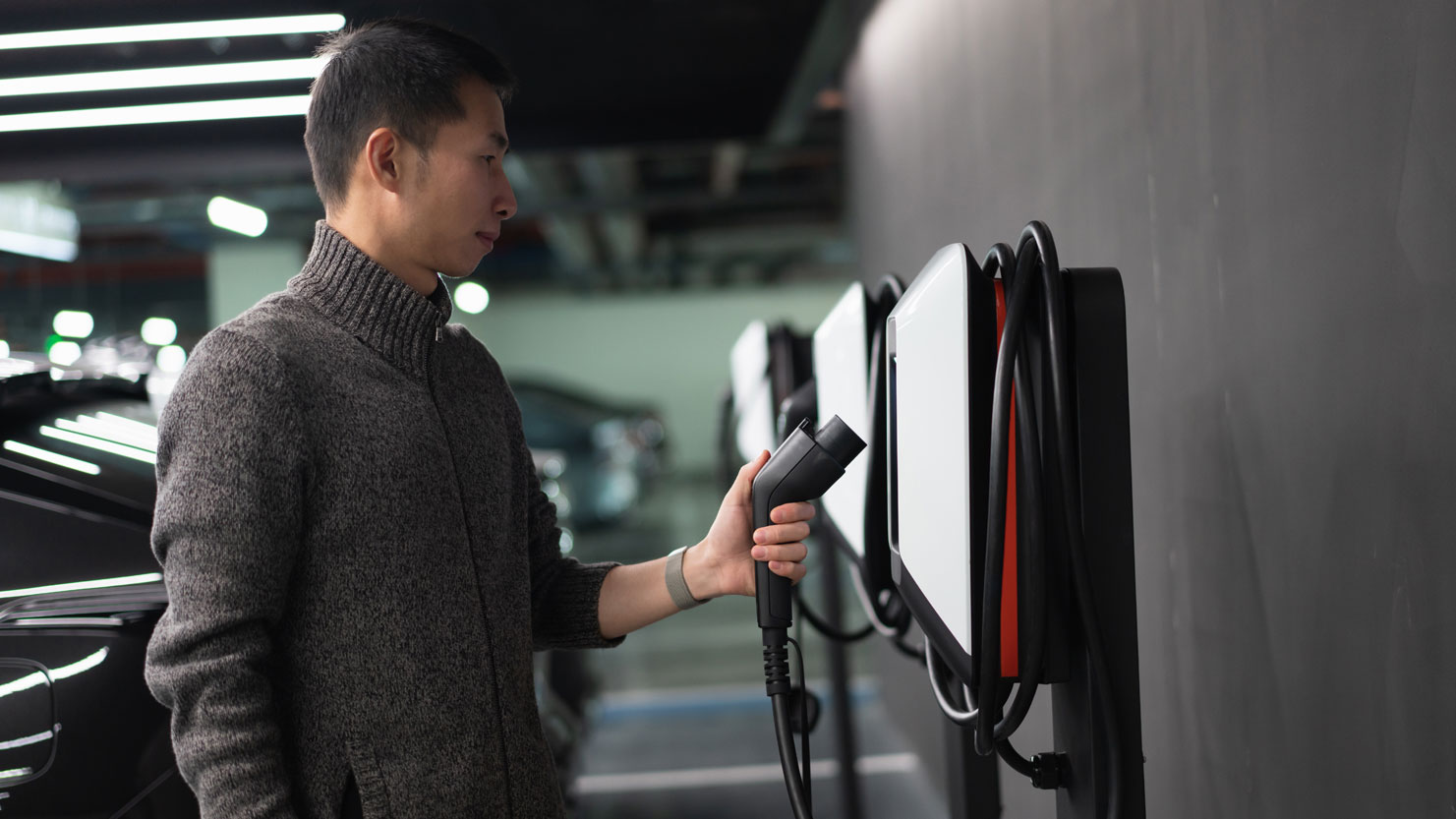 Man standing at a charging station for electric vehicles in a parking garage, holding a charging plug in his left hand. In the background, several cars parked in a row. Man standing at a charging station for electric vehicles in a parking garage, holding a charging plug in his left hand. In the background, several cars parked in a row.