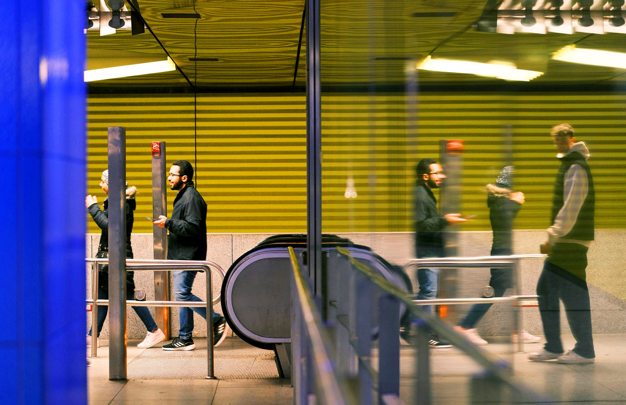 The picture shows a cross-section of a railway staircase with an escalator. On the left are two people who have just arrived at the top, one of them holding a mobile phone. On the right is a glass reflection in which another person can be seen. The picture shows a cross-section of a railway staircase with an escalator. On the left are two people who have just arrived at the top, one of them holding a mobile phone. On the right is a glass reflection in which another person can be seen.